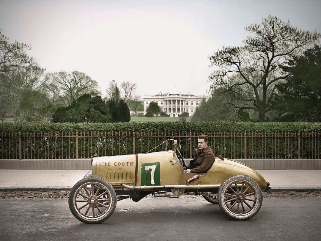 A young man sits in a vintage yellow race car labeled "THE COOTIE" and number 7, parked on a street with the White House visible in the background behind an iron fence and trees.