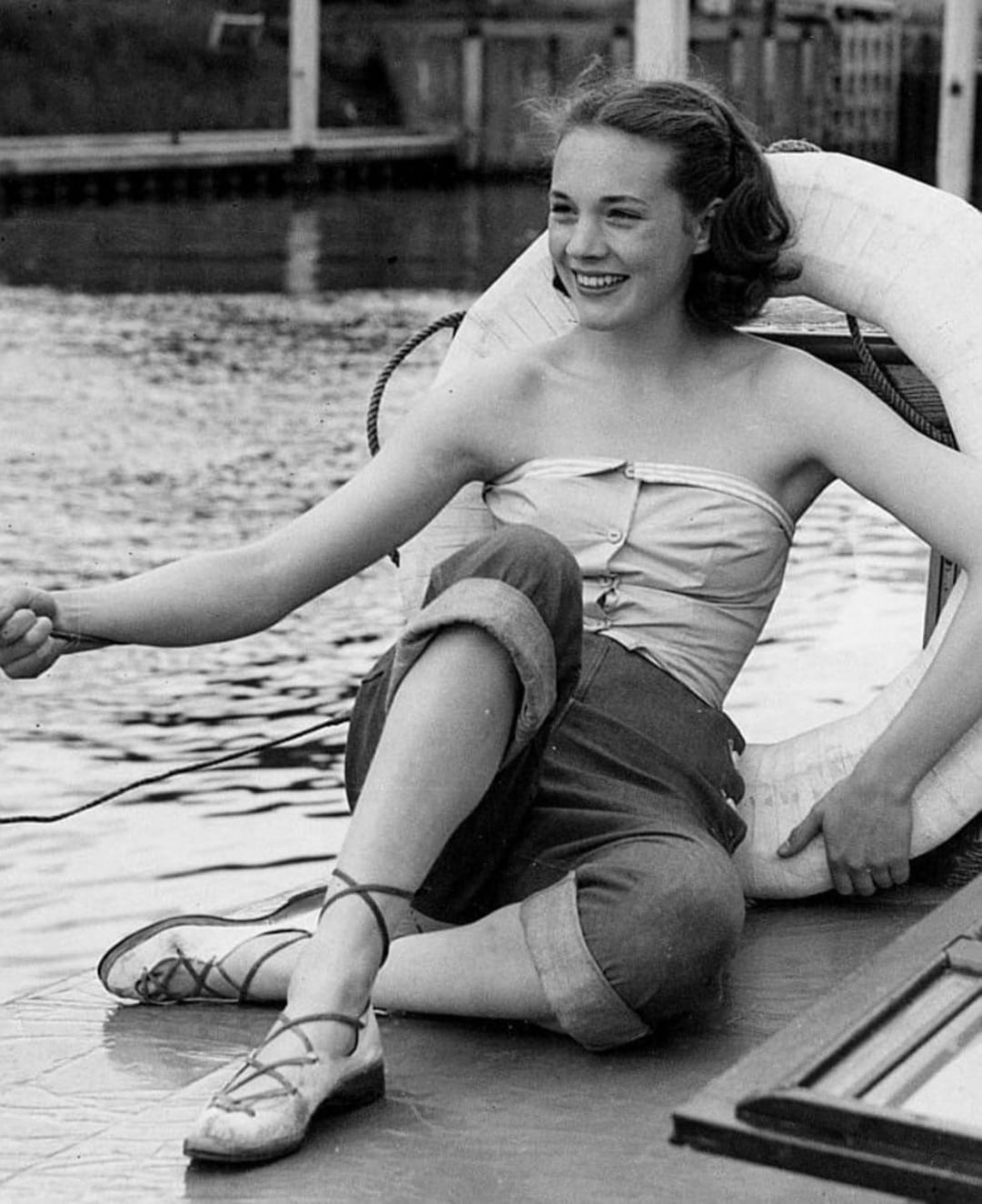 A young woman sits on a dock by the water, smiling and holding a rope. She wears capri pants, a sleeveless top, and flat lace-up shoes. There is a large life preserver behind her and a wooden railing in the background.
