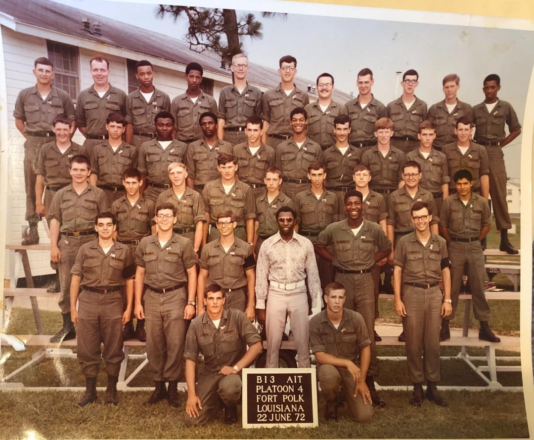 A group of young men in military uniforms pose on bleachers outside a building. One man in light-colored civilian clothes stands in front. A sign reads: “B13 AIT Platoon 4 Fort Polk Louisiana 22 June 72.”