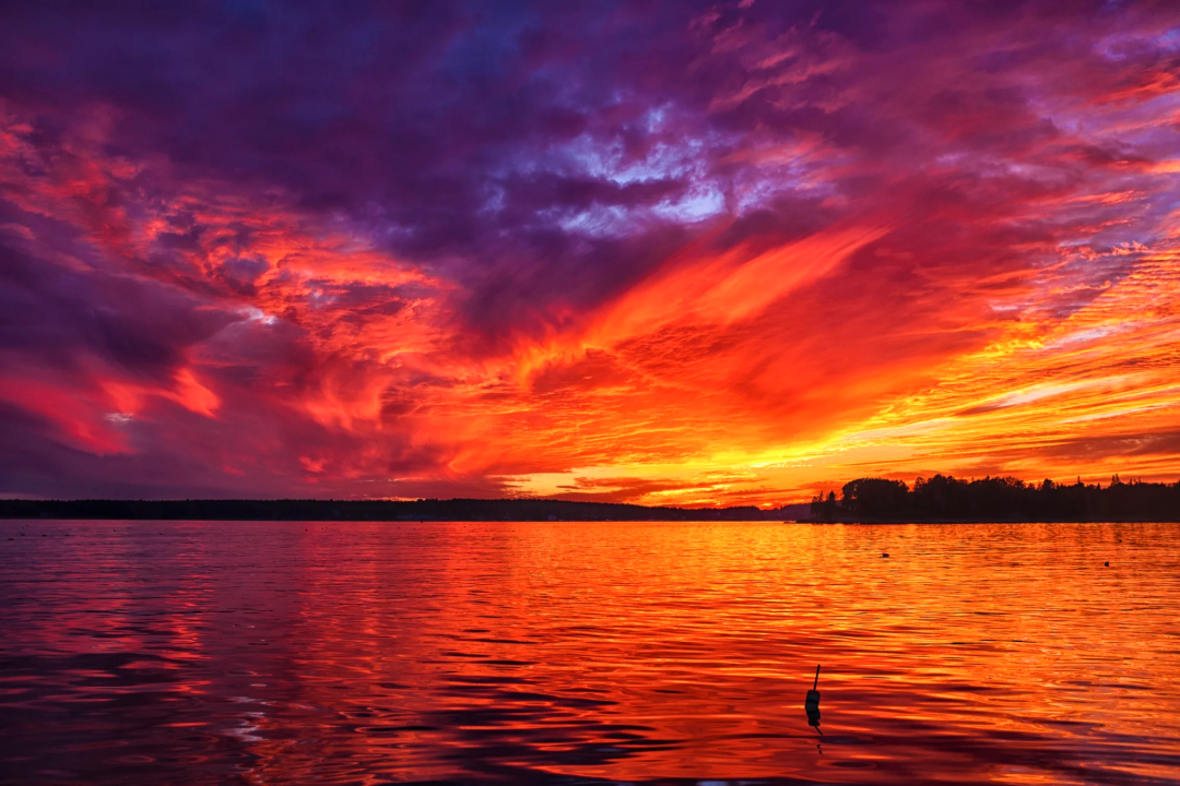 A vibrant sunset over a calm lake, with dramatic clouds painted in shades of orange, red, purple, and blue. The vivid colors are reflected on the water’s surface, and a dark tree line is visible on the horizon.