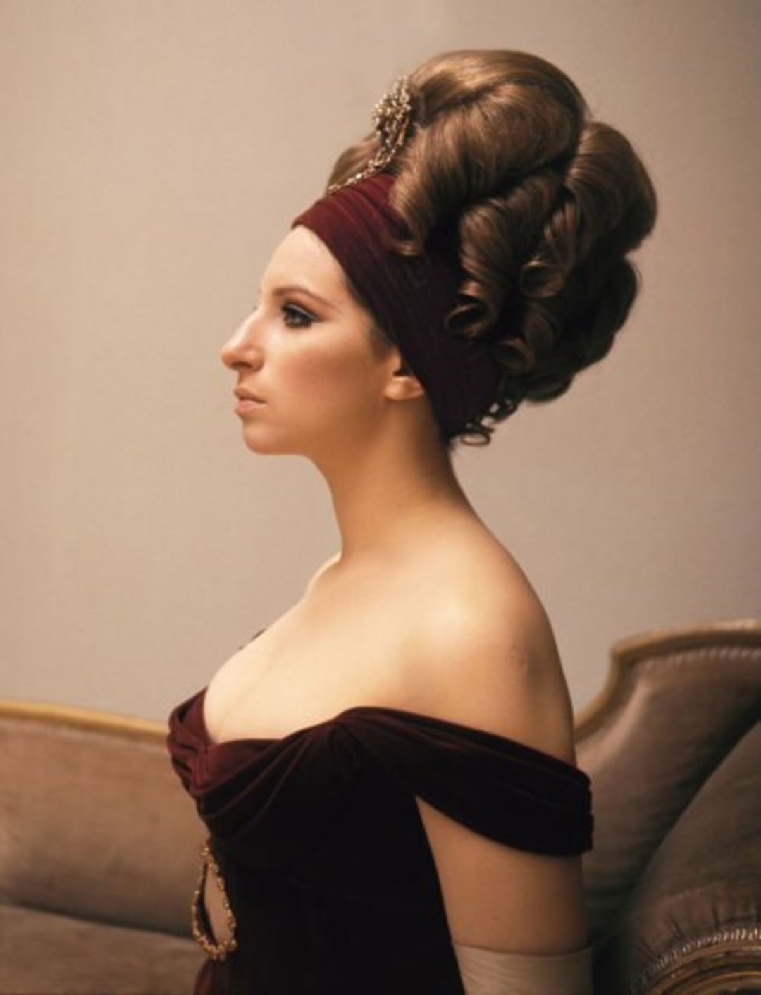 A woman in profile wears an elaborate updo with a jeweled hairpiece and a burgundy headband. She has on an off-the-shoulder burgundy dress and sits on an upholstered chair against a neutral background.