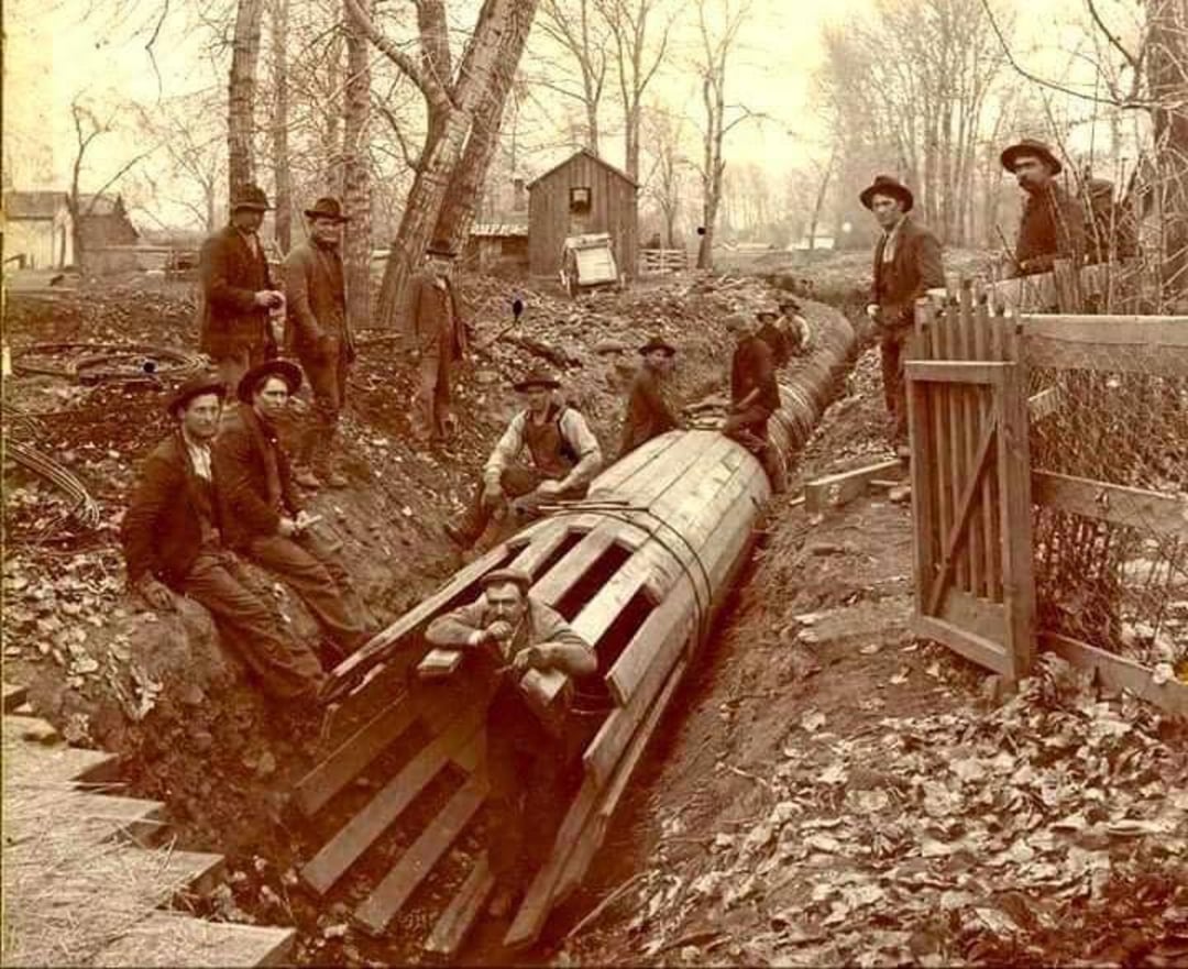 Sepia photo of men in old-fashioned clothing working on a large wooden pipe in a trench, surrounded by trees and autumn leaves, with a wooden fence and small shed in the background.