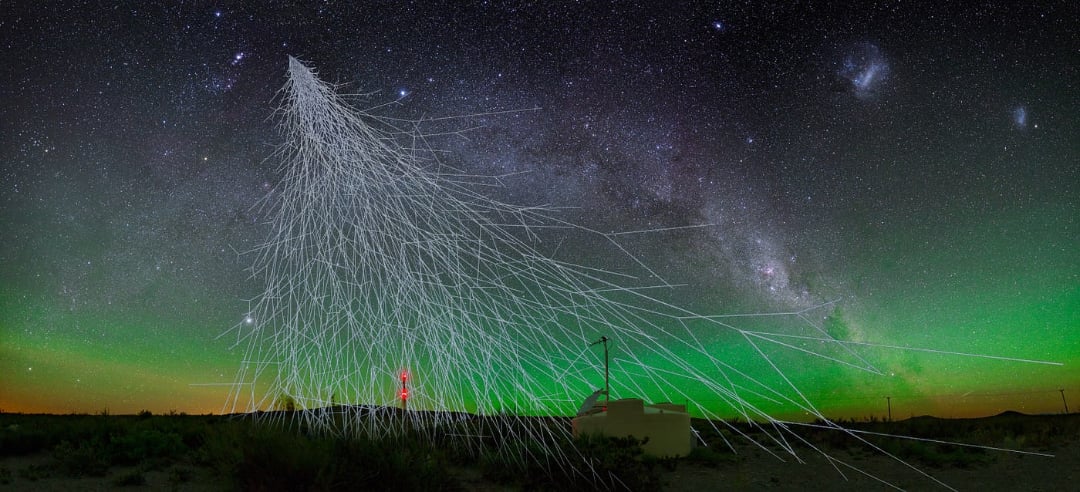 Night sky with green aurora and stars, including the Milky Way. White lines illustrate cosmic ray particle paths descending to a ground-based observatory with scientific equipment and a red-lit antenna on the grassy landscape.