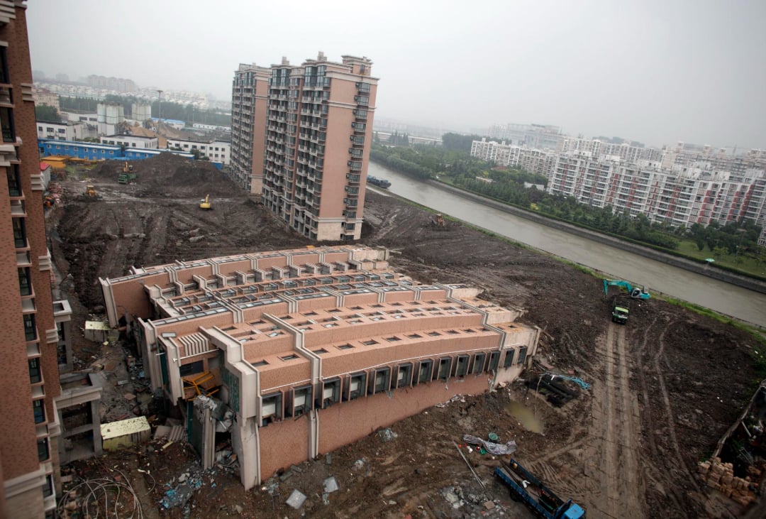 An apartment building in China leans heavily to one side on muddy ground, partially collapsed, with construction vehicles nearby and other intact buildings and a river in the background under a cloudy sky.
