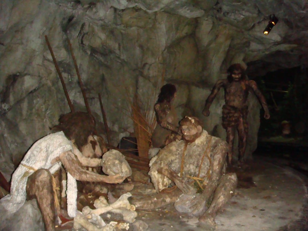 Four prehistoric human figures are depicted inside a cave, surrounded by rocks, bones, and primitive tools, resembling a scene of early human life or a diorama in a museum exhibit.