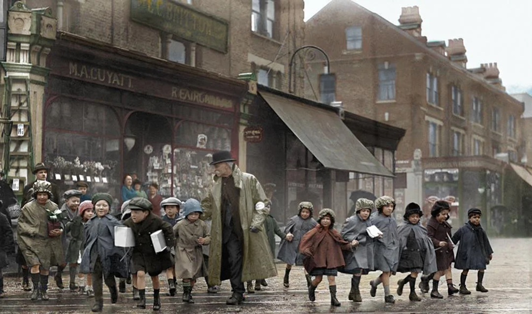 A group of young children in coats, led by a man in a trench coat and hat, cross a wet city street in front of old brick buildings and shops on a rainy day.