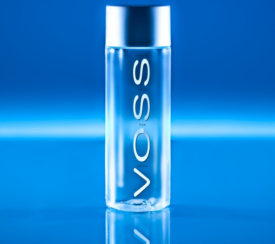 A clear Voss water bottle with a silver cap stands upright on a reflective blue surface, with the Voss logo vertically displayed on the bottle against a blue background.