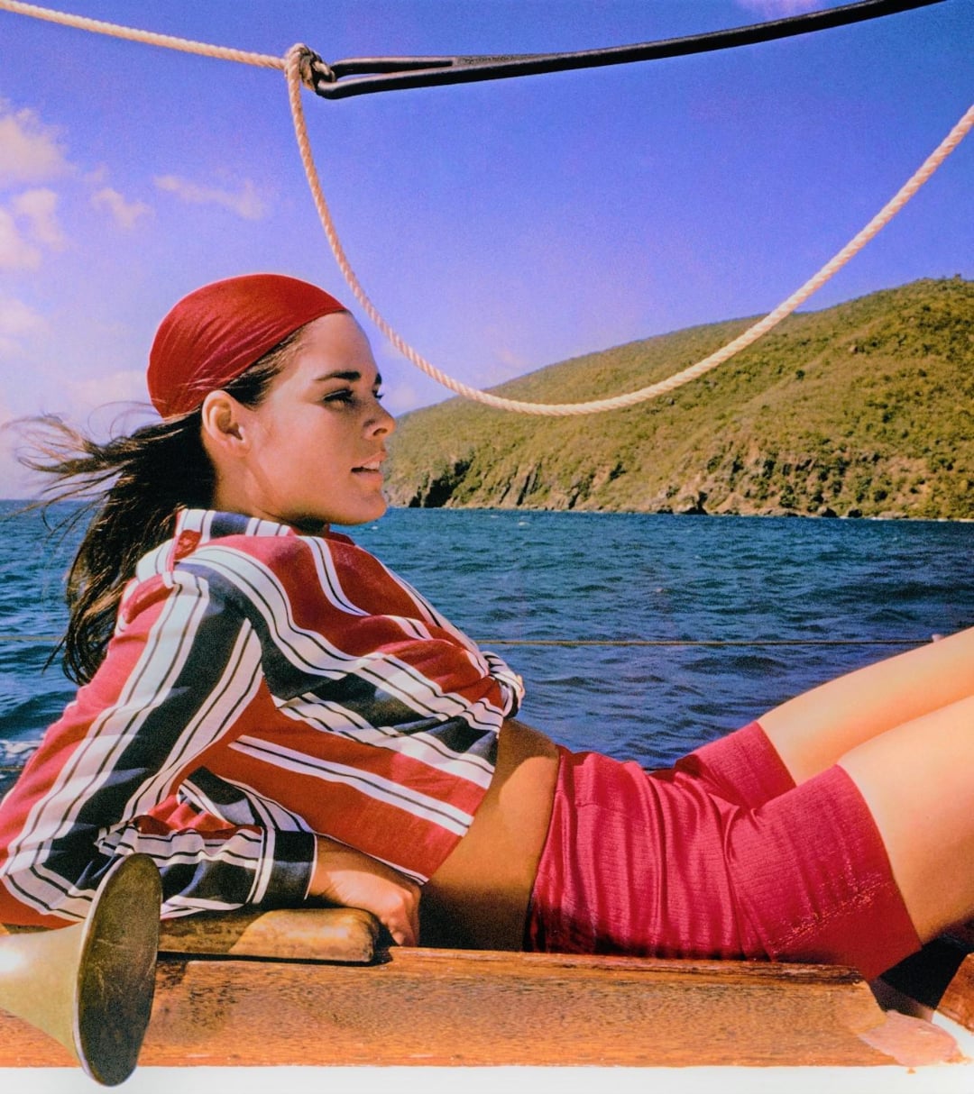 A woman wearing a red headscarf, striped shirt, and red shorts reclines on a boat, gazing toward the shore, with blue sea and green hills in the background under a clear sky.