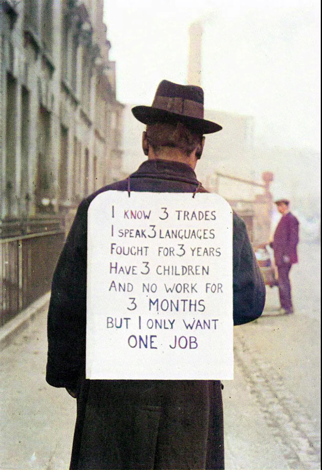 A man in a coat and hat stands with his back to the camera, wearing a sign that says he knows three trades, speaks three languages, fought for three years, has three children, and seeks one job after three months without work.