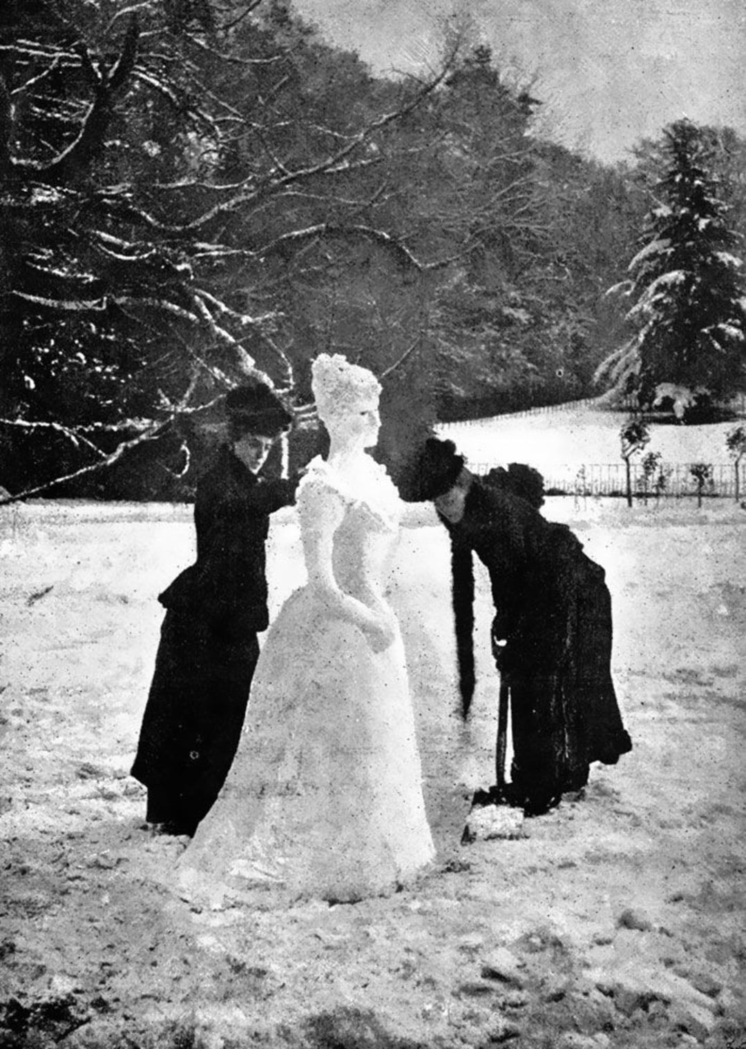 Two women in dark Victorian-era clothing stand in the snow, shaping a life-sized snowwoman. Bare trees and a snow-covered landscape are visible in the background.