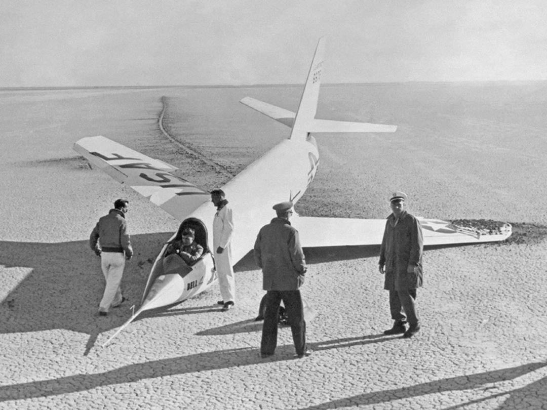 Four men stand near a large glider with “712” on the tail, on a cracked, dry lakebed. One man sits in the cockpit, while the others examine the aircraft and the surrounding area.