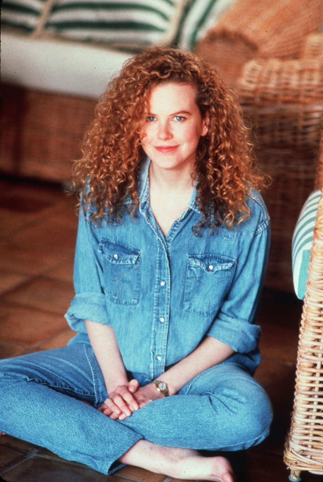 A woman with long curly red hair sits cross-legged on the floor, wearing a denim shirt and jeans. She is smiling softly and is surrounded by wicker furniture with striped cushions.
