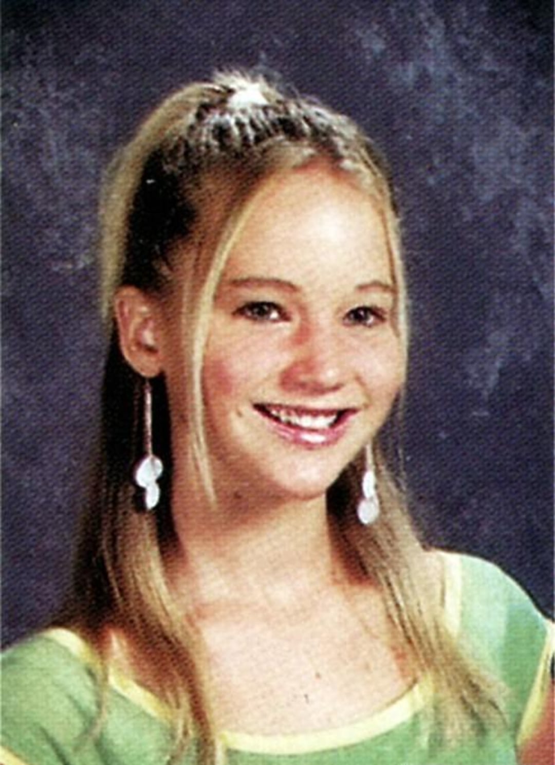A teenage girl with long blonde hair in a high ponytail smiles at the camera. She wears dangling earrings and a green top with yellow trim, posed against a mottled, dark gray school portrait background.