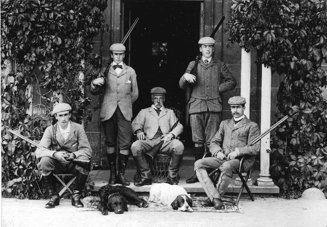 Five men in vintage hunting attire pose with rifles outside a building, flanked by ivy. Two men stand, three are seated, and two dogs lie on the ground in front of them. The scene appears early 20th century.