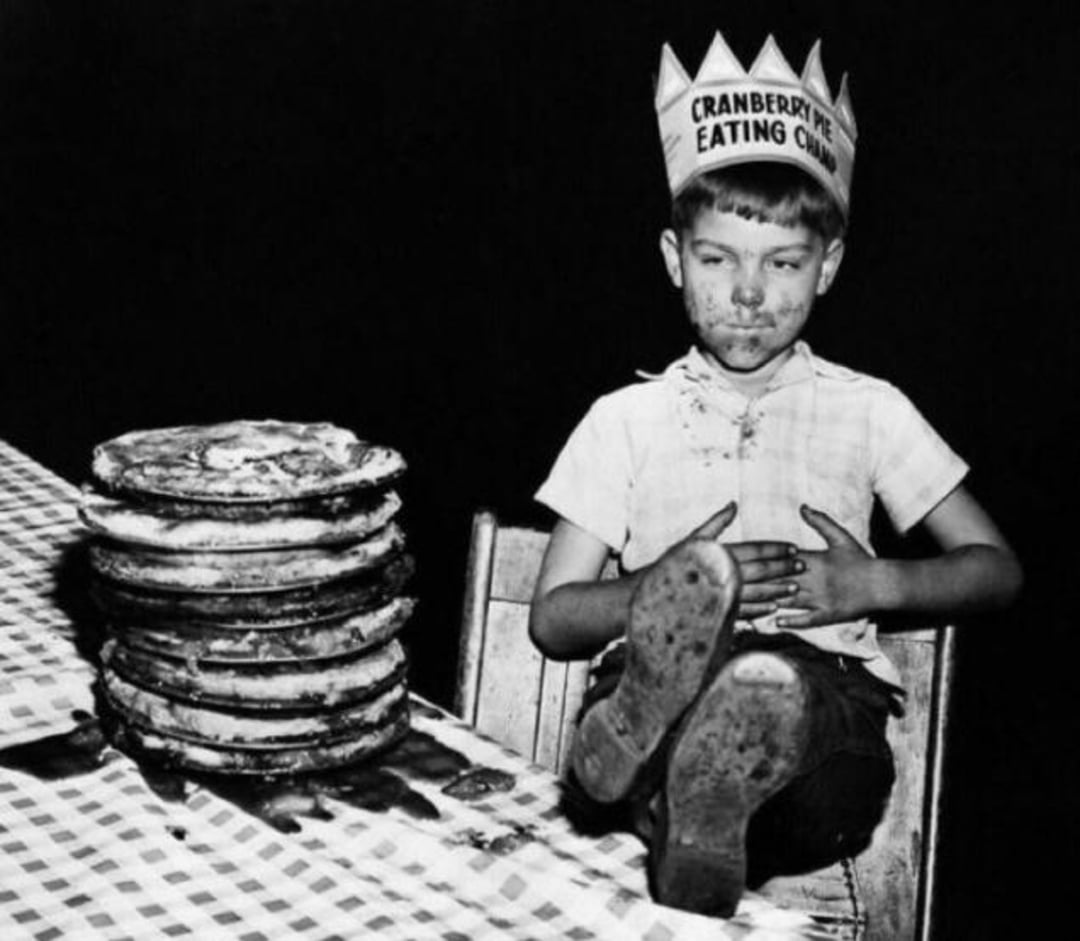 A boy wearing a paper crown labeled "Cranberry Pie Eating Champ" sits back with a full stomach, his face messy, next to a tall stack of empty pie tins on a checkered tablecloth.