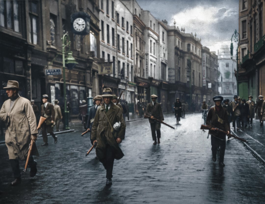 A group of armed men in coats and hats walk down a wet, empty city street lined with old buildings under a cloudy sky. Some carry rifles and wear sashes; a clock hangs on a lamppost in the background.