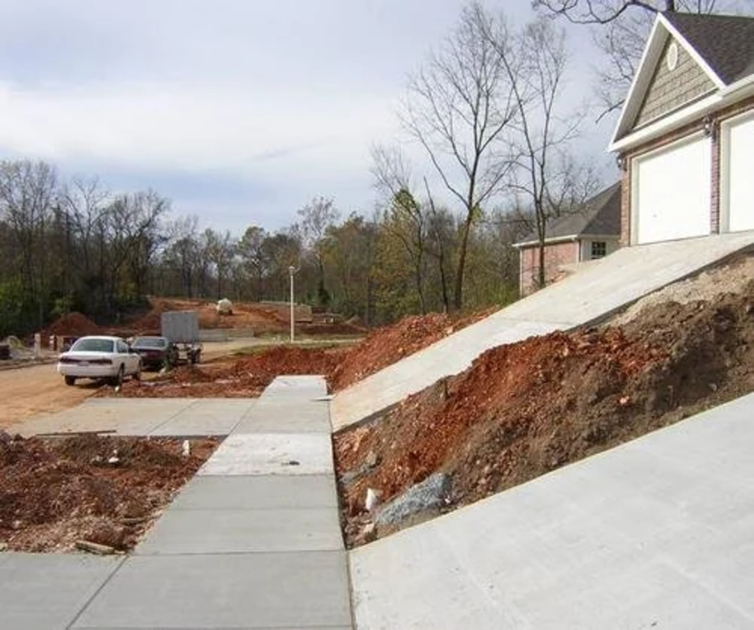 A sloped driveway leads up to a garage above a steep, unfinished dirt embankment. The sidewalk below runs parallel to the slope. In the background, there are trees, a parked car, and construction equipment.