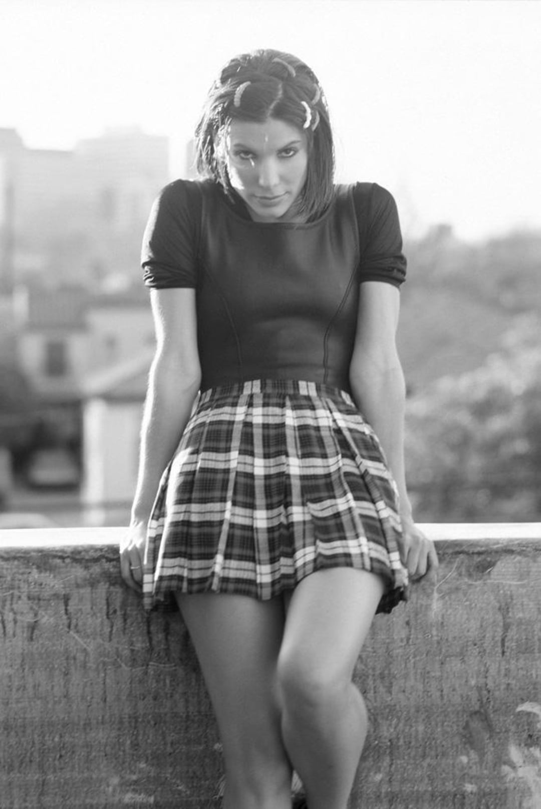 A woman wearing a short-sleeved top and a plaid skirt leans against a concrete ledge outdoors. She looks directly at the camera with a serious expression. The photo is in black and white.