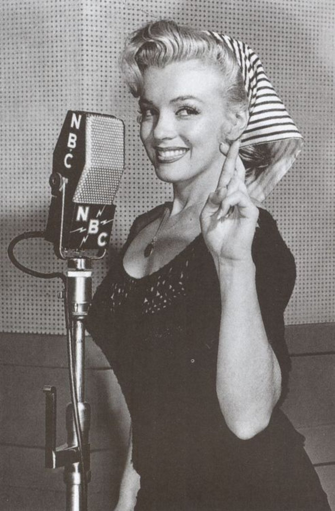 A woman with curled blonde hair and a striped headscarf stands in front of a vintage NBC microphone, smiling and holding her fingers crossed. The image is in black and white.