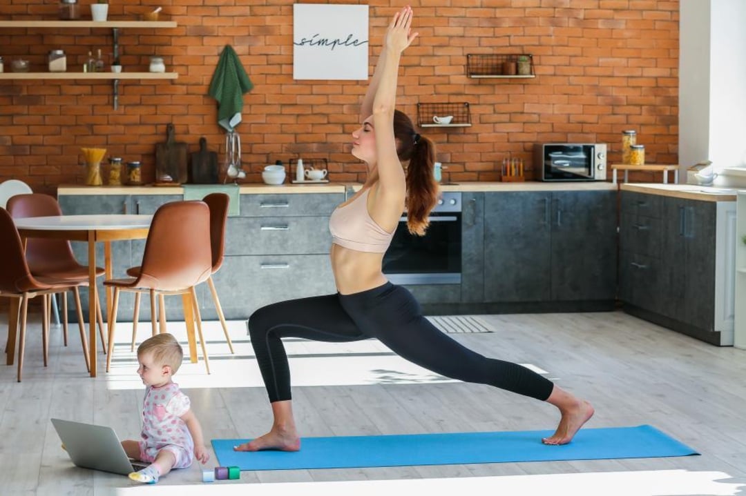 A woman practices yoga on a mat in a modern kitchen while a baby sits nearby on the floor looking at a laptop.
