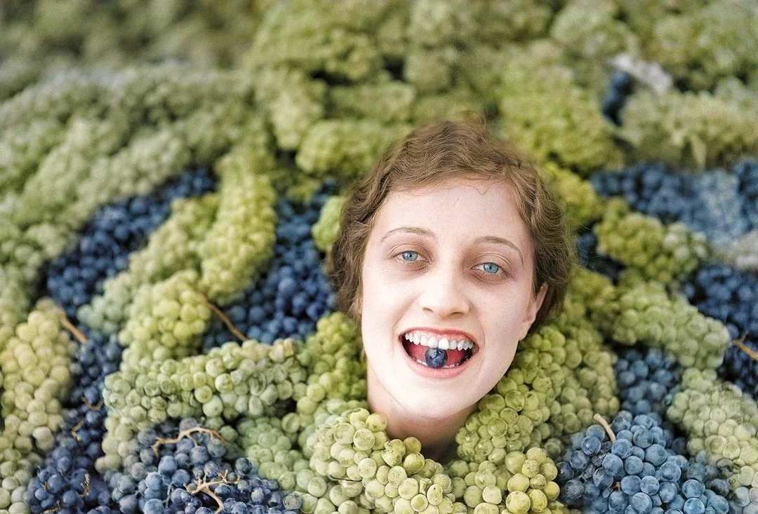 A smiling woman’s head emerges from piles of green and purple grapes, with grapes held between her teeth. The background is entirely covered in bunches of grapes.
