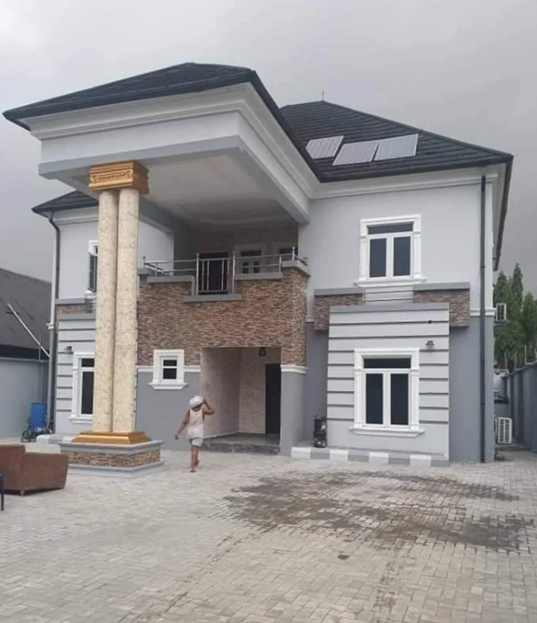 A two-story modern house with gray walls, white trim, brick accents, and large columns in front. A person in white clothes stands in the paved driveway. The sky is overcast.
