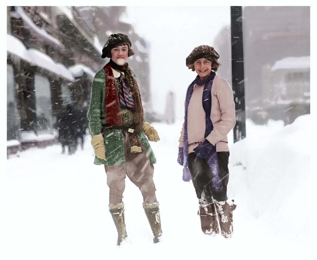 Two people wearing winter clothes and hats stand smiling in a snow-covered city street, surrounded by deep snow and falling snowflakes. Buildings and a distant figure are visible in the blurry background.