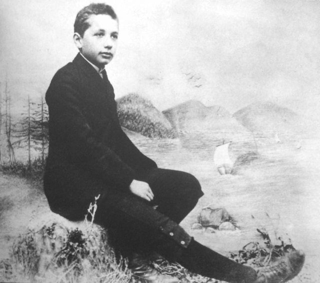 A young boy in a dark suit sits on a rock with a painted backdrop of mountains, water, and sailboats behind him. The photo is black and white and appears to be from the late 19th or early 20th century.