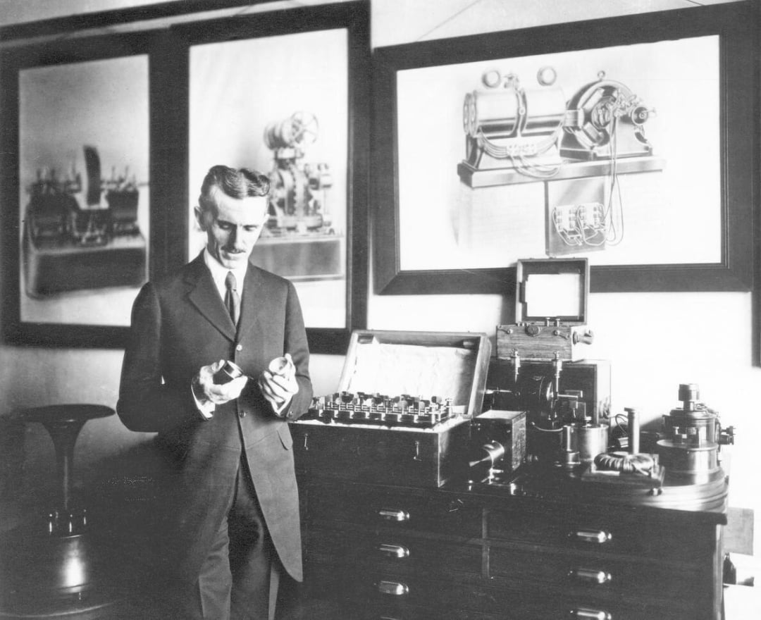 A man in a suit stands in a room holding two objects, surrounded by electrical equipment on a table. Large framed technical drawings and photos of machines hang on the wall behind him. The scene appears historical.
