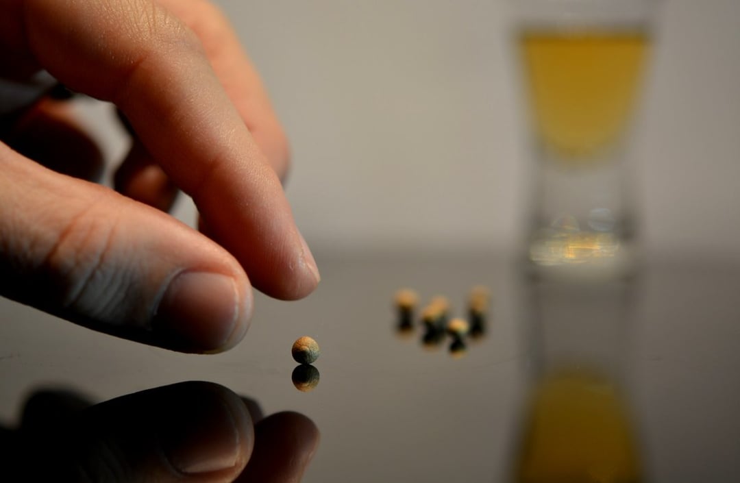 A close-up of a hand picking up a small round object from a reflective surface, with several similar objects nearby. In the blurred background, there is a small glass containing a yellow liquid.