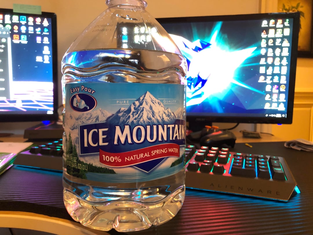 A large bottle of Ice Mountain spring water sits on a desk in front of two computer monitors with colorful screensavers and a backlit Alienware keyboard.