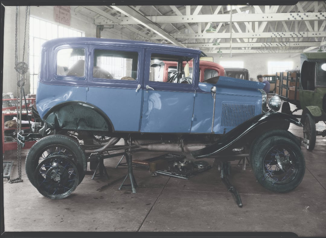 A vintage blue car is elevated on a workshop floor, its wheels removed and undercarriage exposed. The garage is filled with tools and other classic cars, and sunlight streams through large windows.