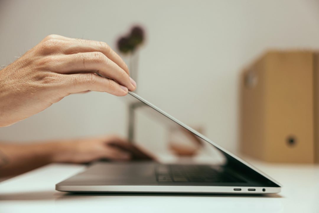 A person’s hand is partially closing a slim laptop on a white desk, with a blurred hand in the background and a cardboard box and a plant visible on the right.
