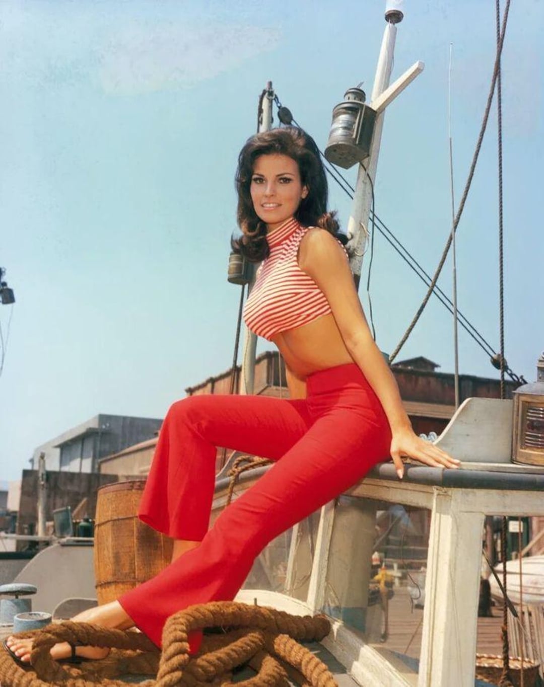 A woman with dark hair in a red and white striped crop top and red pants sits on the deck of a boat, smiling. Nautical equipment and buildings are visible in the background under a clear blue sky.