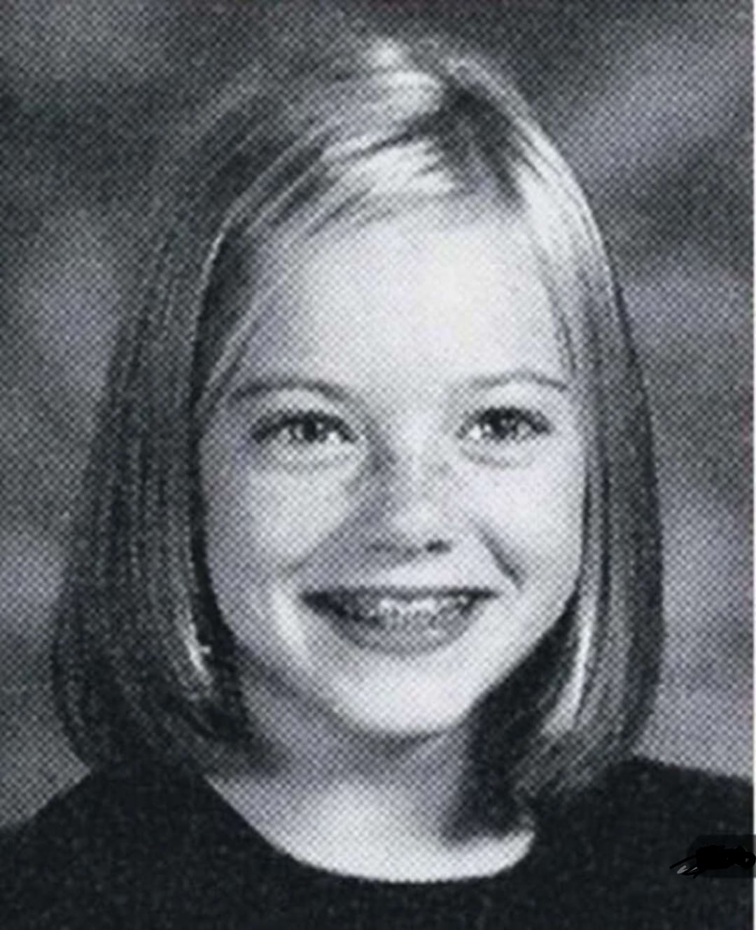 A black-and-white yearbook photo of a young girl with straight, shoulder-length hair, wearing a dark top and smiling at the camera. The background is softly blurred.