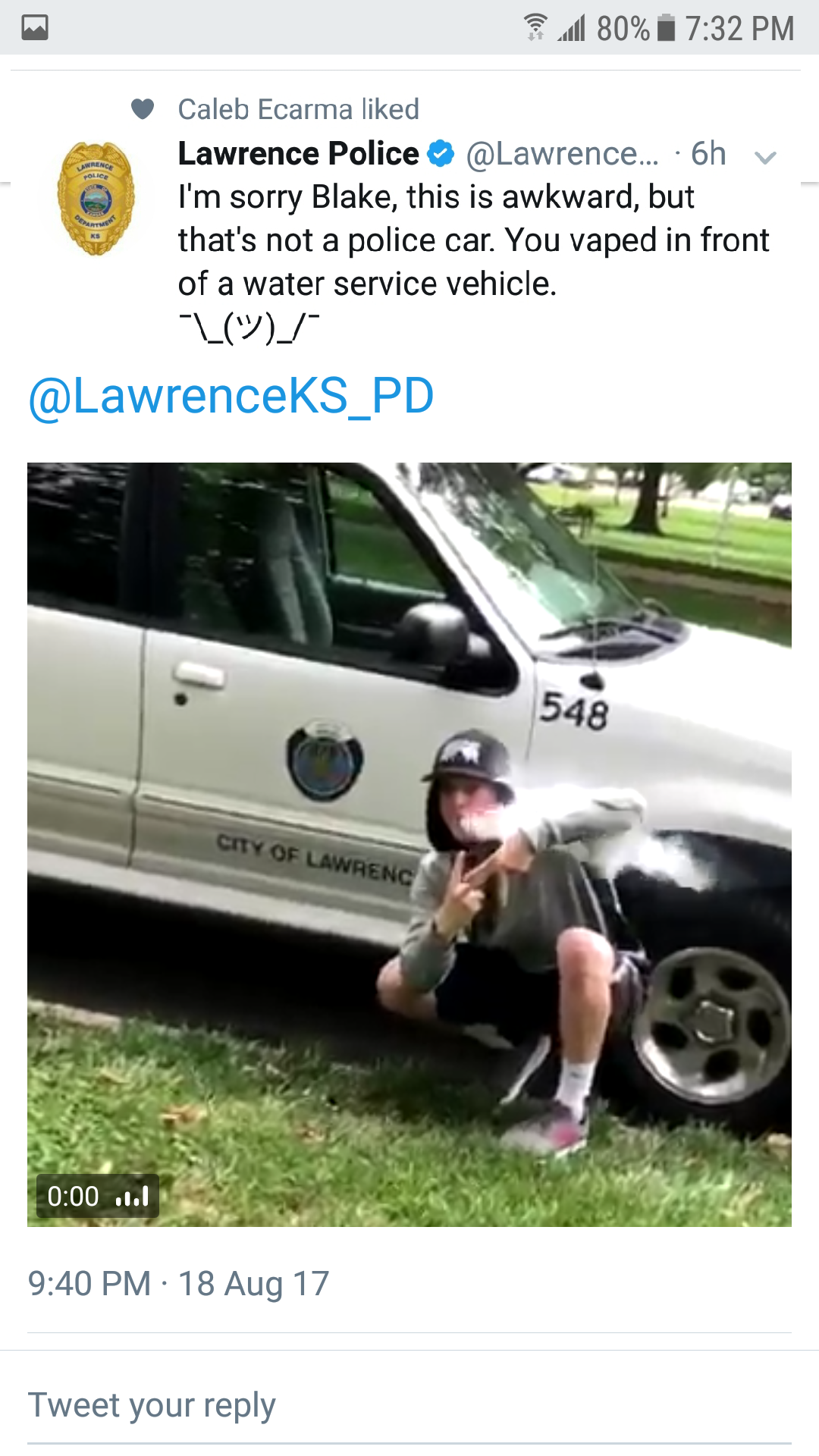 A young person squats and poses in front of a white City of Lawrence water service vehicle, wearing a hat, hoodie, and shorts. The tweet above jokes about mistaking the vehicle for a police car.