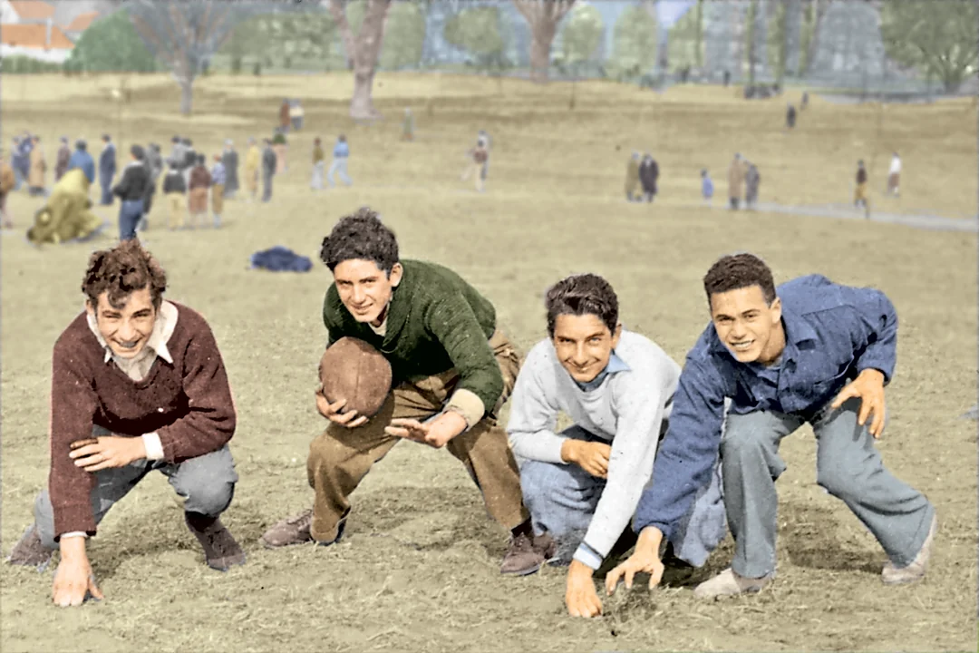 Four teenage boys pose in a crouched football stance on a dirt field, one holding a ball. People are scattered across the field in the background on a bright day.