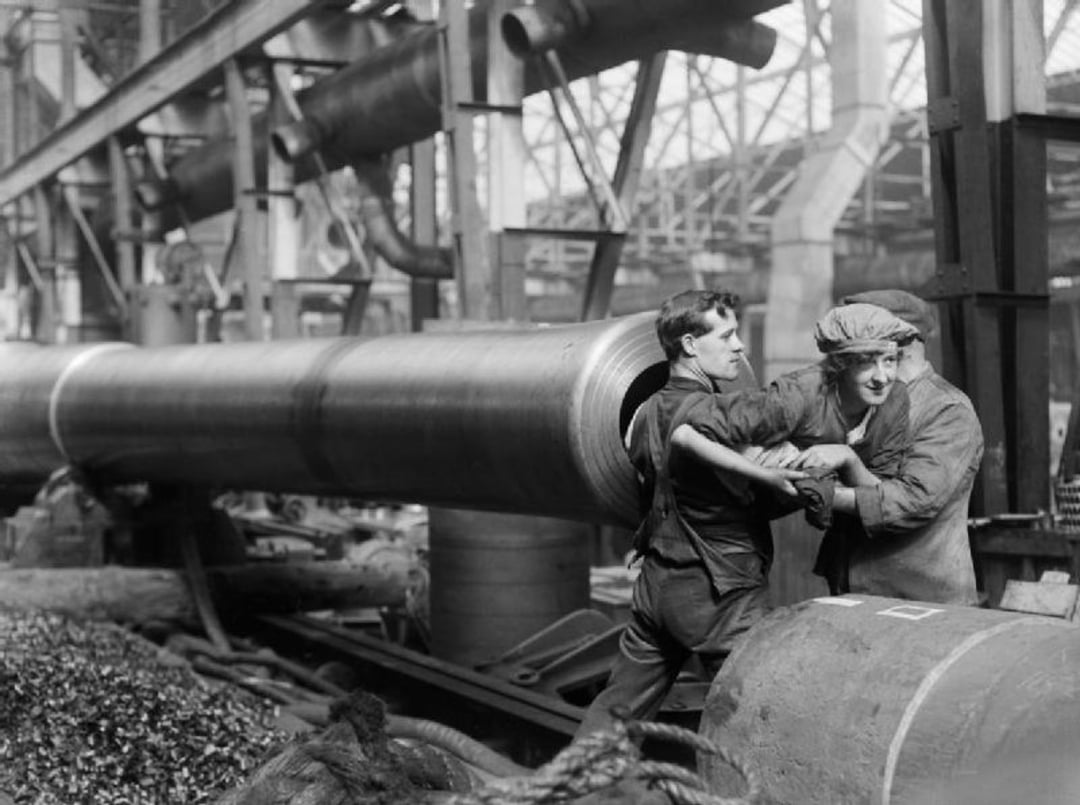 Two workers in overalls and caps smiling and playfully interacting while working with large metal pipes in an industrial factory setting filled with machinery and equipment.