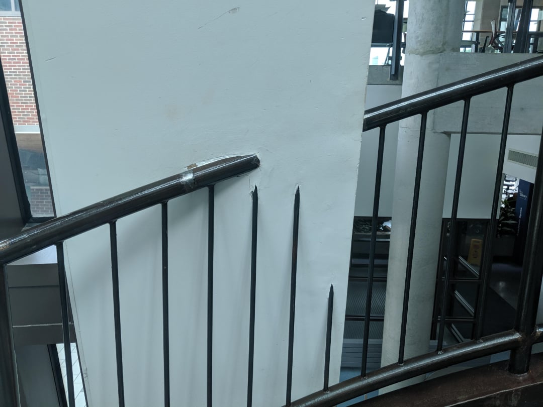 A staircase railing curves abruptly into a white wall, with the metal bars piercing and bending into the plaster, creating a visibly awkward and damaged intersection. Natural light enters through nearby windows.