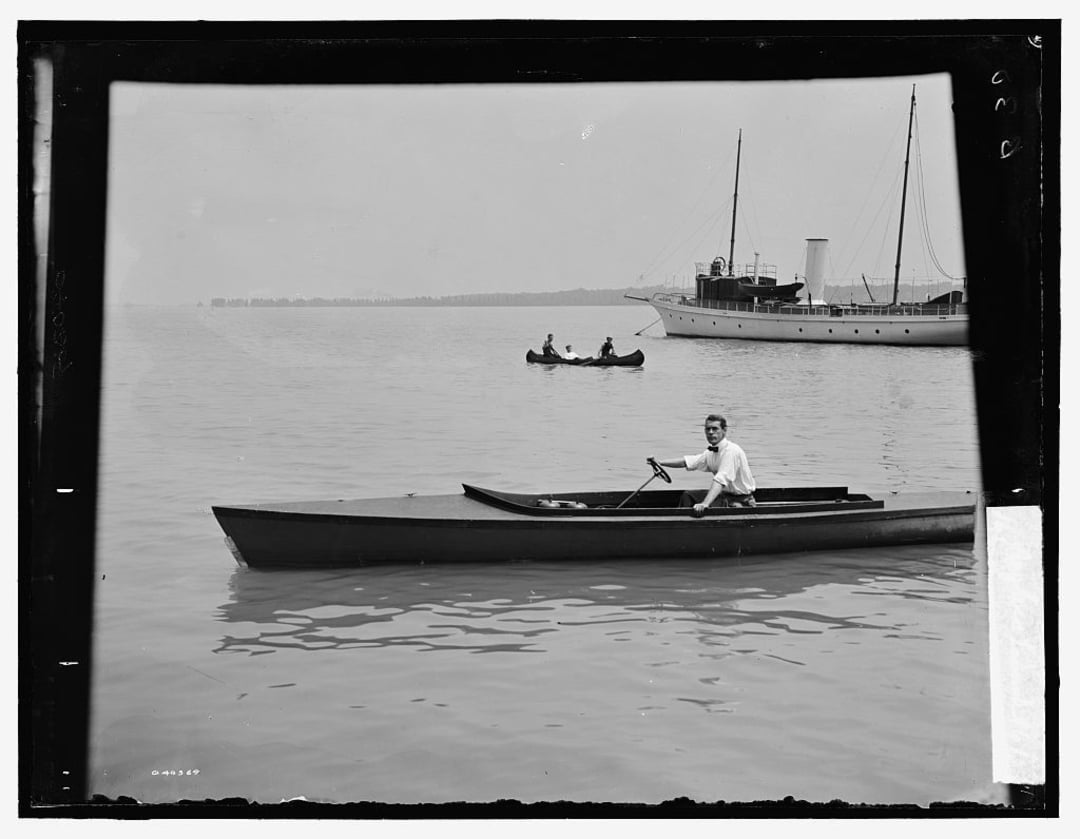 A man rows a small wooden boat on calm water, with a steamship and a canoe with two people in the background under a hazy sky.