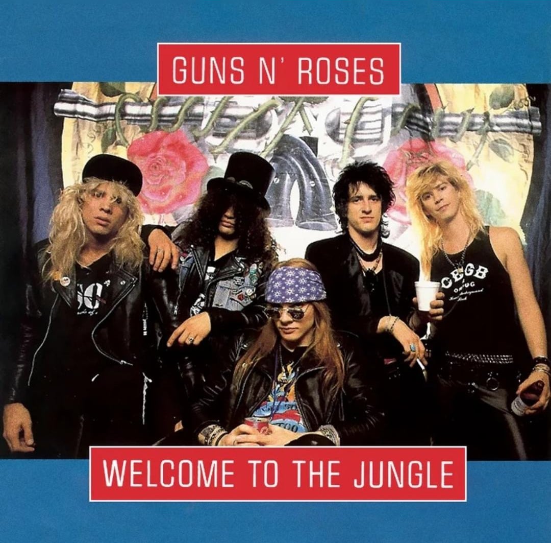 Five members of Guns N’ Roses pose together indoors, dressed in rock attire. Above them, "Guns N’ Roses" is written in red on a blue background. Below, “Welcome to the Jungle” is displayed in white on red.