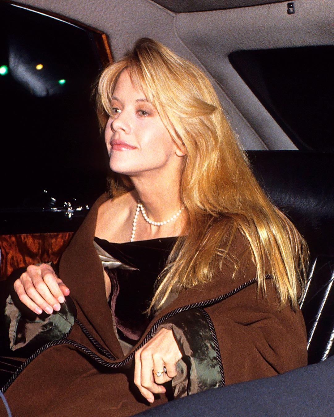 A woman with long blonde hair, wearing a brown shawl and a pearl necklace, sits in the backseat of a car, looking out the window.