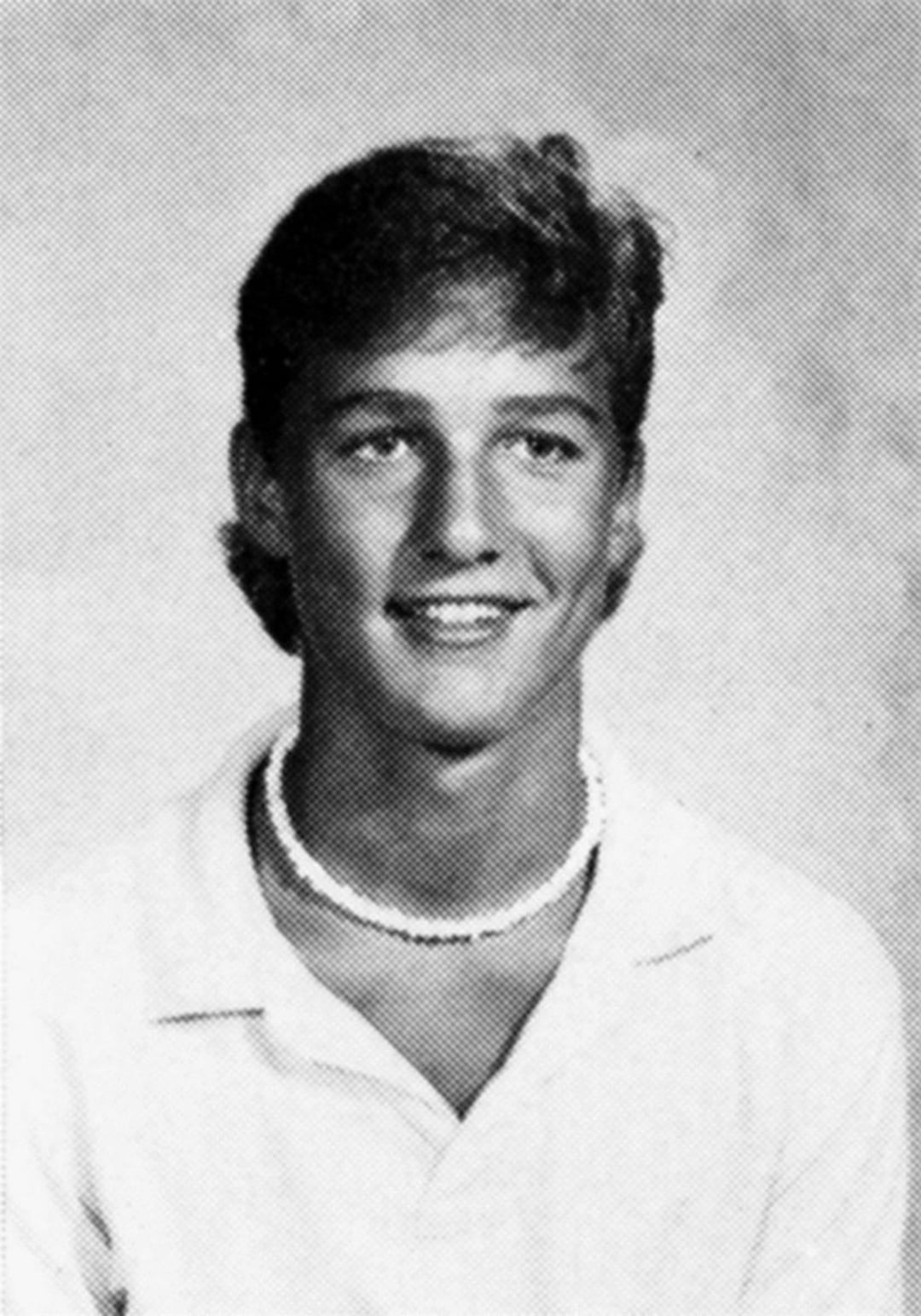 Black and white yearbook photo of a young man with short hair, wearing a white collared shirt and a beaded necklace, smiling at the camera against a plain background.