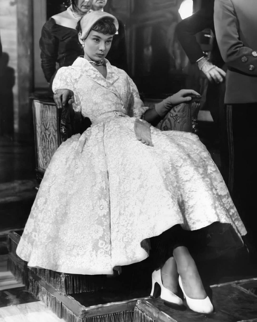 A woman in a vintage, full-skirted dress and headband sits confidently on a chair with one arm resting on the back, in a black-and-white, classic fashion scene. Other people are partially visible in the background.