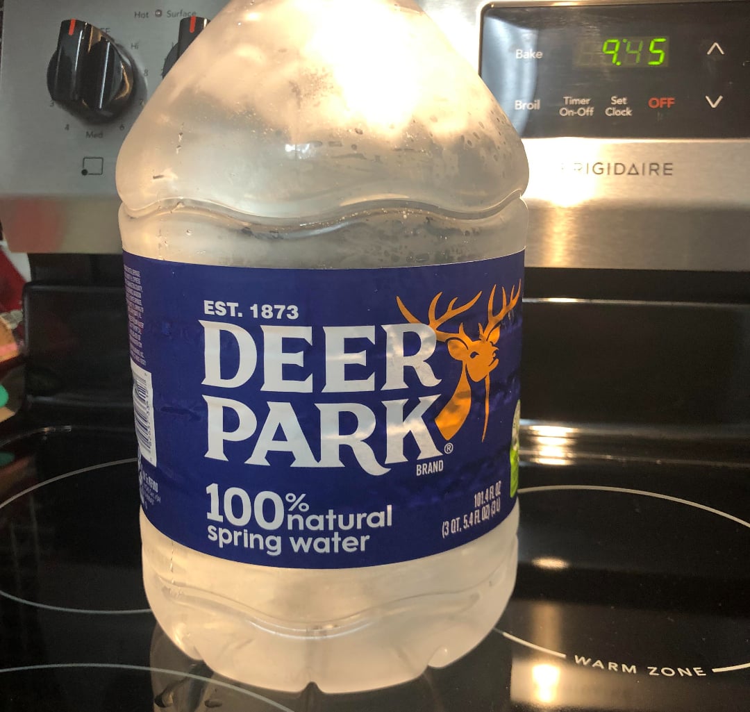 A large plastic jug of Deer Park 100% natural spring water sits on a stovetop. The blue label features a yellow deer head logo. An oven with the digital clock reading 9:45 is in the background.