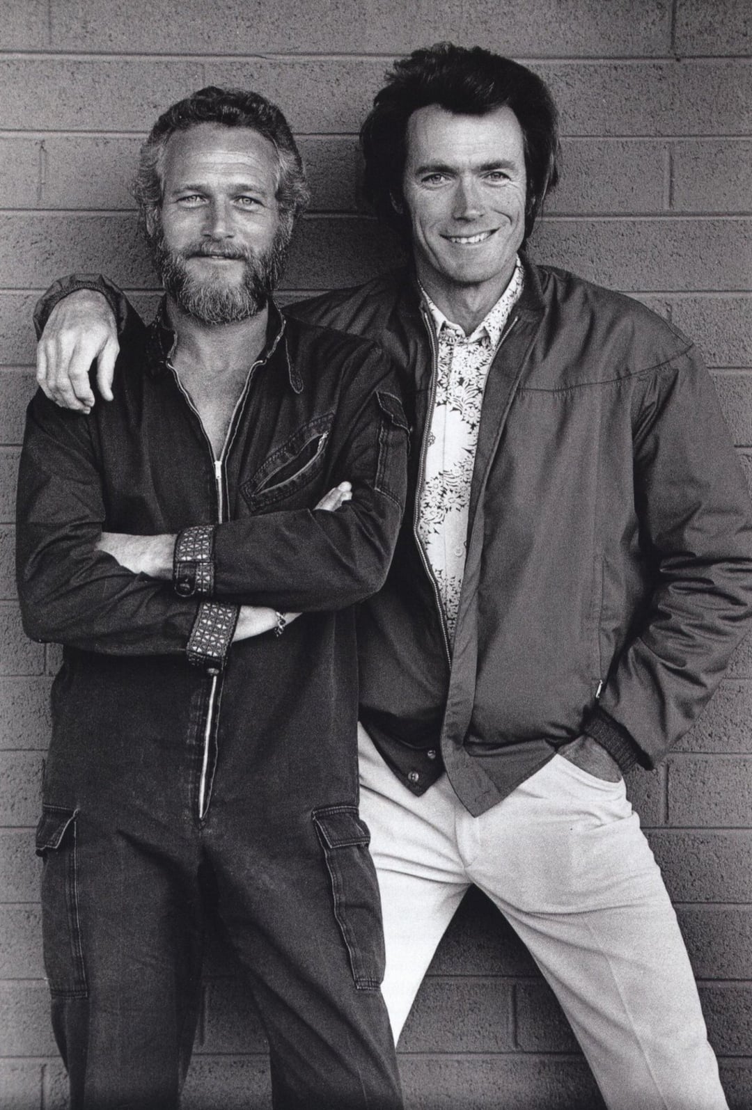 Two men stand smiling against a brick wall. One has a beard and wears a dark jumpsuit with folded arms, while the other is clean-shaven, wearing a light shirt and jacket, with one arm around the other's shoulder.