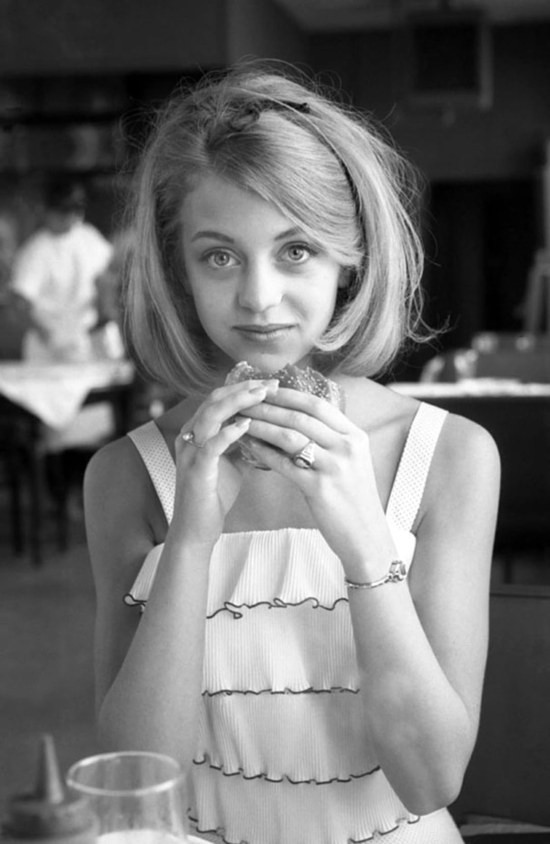 A young woman with shoulder-length blonde hair and a sleeveless, ruffled dress sits at a table in a restaurant, holding a sandwich with both hands and looking directly at the camera. The image is black and white.