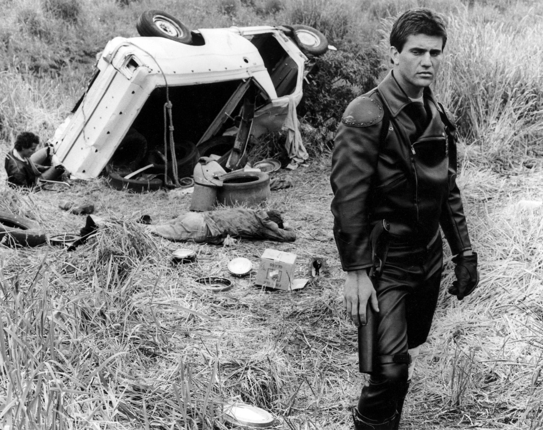 A man in black leather stands in the foreground of a grassy field, looking serious. In the background, an overturned vehicle and debris are scattered, with two people lying on the ground near the wreck.