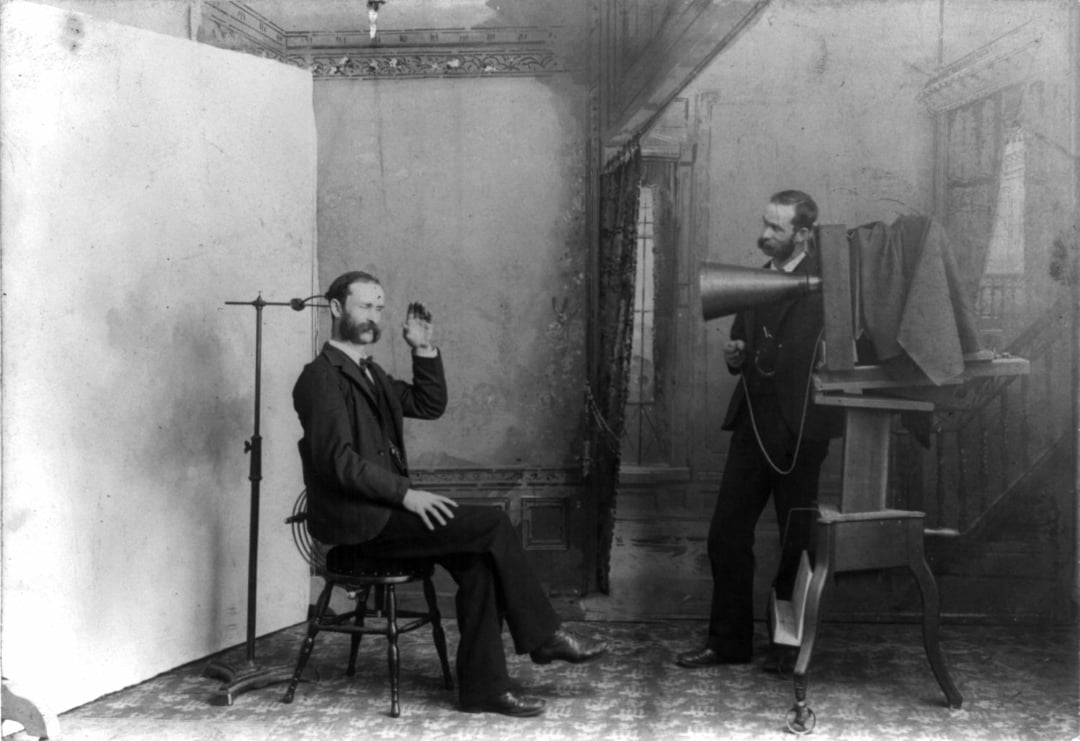 A vintage black-and-white photo shows a man sitting on a chair, speaking or waving, while another man stands behind a large early recording device with a horn in a decorated studio room.