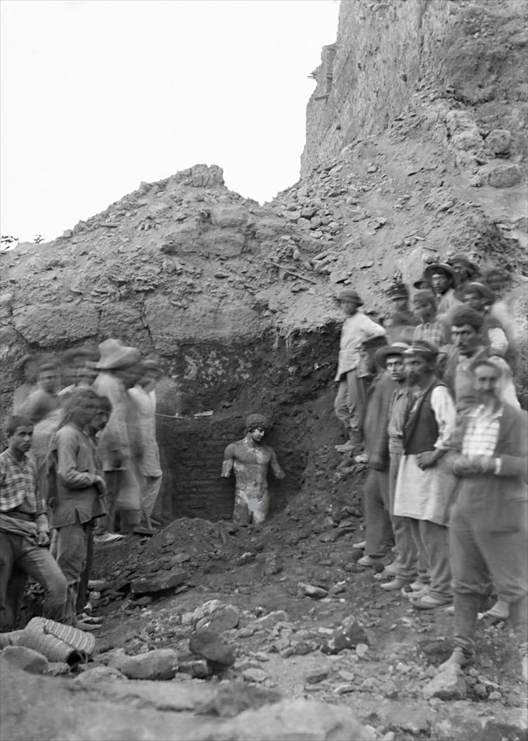 A group of people stand around an excavation site where a partially unearthed ancient statue is embedded in soil, with rocky terrain and a steep dirt mound in the background.